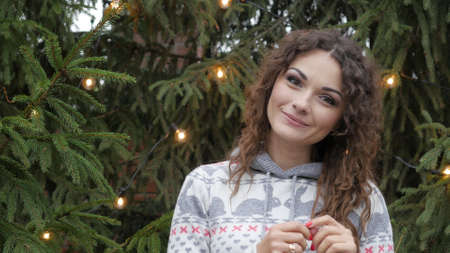 Cheerful woman in Santa hat with red scarf and mittens is happy and jumping against the background of Christmas tree in garlandsの写真素材