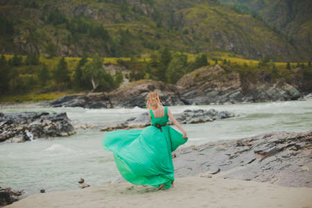 young woman in a long flowing dress on the background of the river and mountains. coloring women goes in a green dress on a background of a beautiful landscape. a woman walks beside a river. soft light.の写真素材