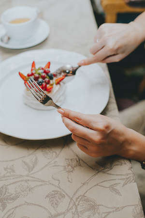 Photo of female hands cutting a fruit cake with strawberries, mint and black currants on a white plate on a table in a restaurant. dessert pavlova. table etiquetteの写真素材