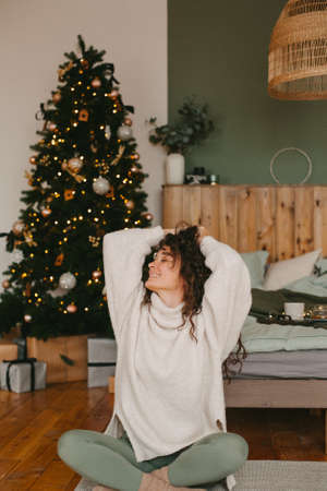 A young woman in cozy home clothes poses on a bed in a loft interior near a Christmas tree.の写真素材