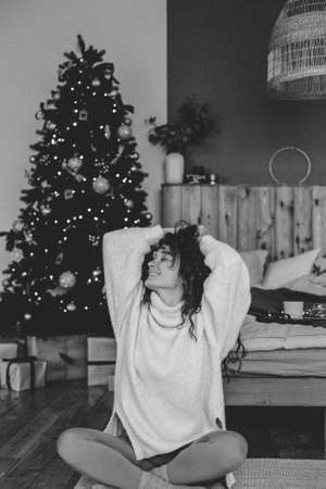 A young woman in cozy home clothes poses on a bed in a loft interior near a Christmas tree.の写真素材