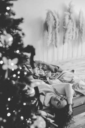 A young woman in cozy home clothes poses on a bed in a loft interior near a Christmas tree.の写真素材