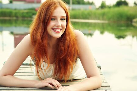 summer portrait of a young redhead woman on background of water in countryside.