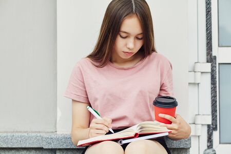 Girl writing pen in notebook. Student with coffee in a paper cup near university.の写真素材