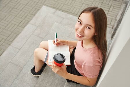 school girl writing pen in notebook on the knees. Student with coffee in a paper cup near university.の写真素材