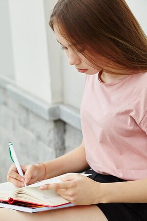 school girl writing pen in notebook on the knees. Student near university.の写真素材