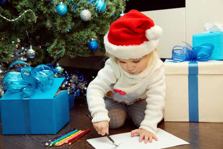 Cute little girl in santa hat writing a letter to Santa Claus near Christmas treeの写真素材