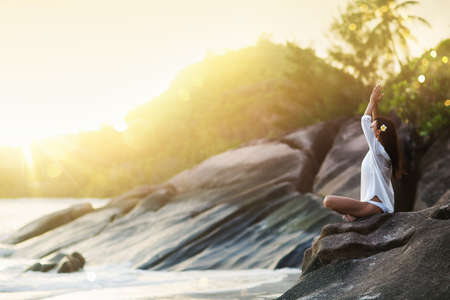 Woman Yoga Meditates on a Rock on the Ocean Beach in the Sunlightの写真素材