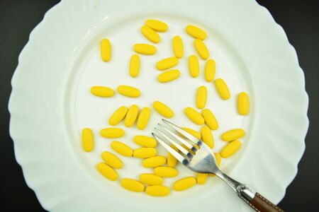 Yellow pills lying on a white plate with silver fork.  Medical overuse, tablet addictionの写真素材