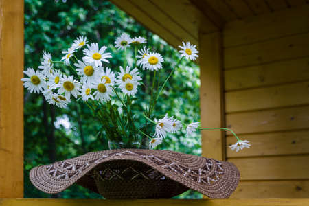 Wicker brown hat and bouquet of daisies in sunny day. Straw cap with chamomiles on rustic wooden terrace. Still life of bunch of white wildflowers with copy space. Summer vacation rural concept.の写真素材