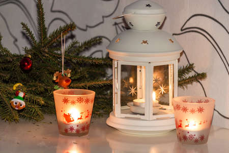 Christmas white hand lantern burning candles with New Year ornaments and spruce branches with reflected on the table on light background. Bright still life interior decor for seasonal winter holidays.の写真素材
