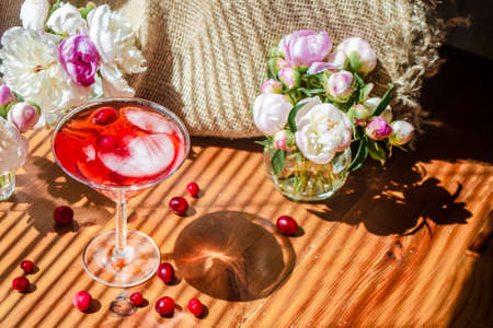 Solemn rustic still life with glass of red drink with autumn berries and ice surrounded by white flowers on textile background. Wooden table in the striped shadow of the blinds on bright sunny day.の写真素材
