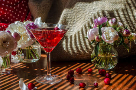 Solemn still life with glass of red drink with autumn berries surrounded by white flowers on a textile background. Wooden table in the diagonal striped shadow of the blinds on a bright sunny day.の写真素材