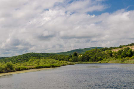 View from the calm river on green trees and bushes against the background of high rocky mountains in the middle of a sunny day. Large white clouds in the sky in summer on a pond. Concept of serenity.の写真素材