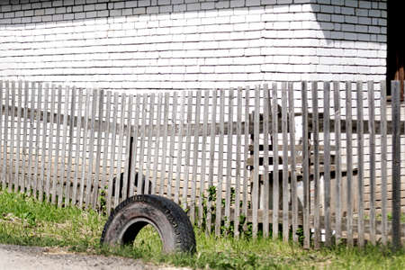 An old grey boardwalk wooden fence with parallel planks and a used tire on the green grass in front of a white brick wall on a bright sunny day. A concept of rural life with space for text.の写真素材