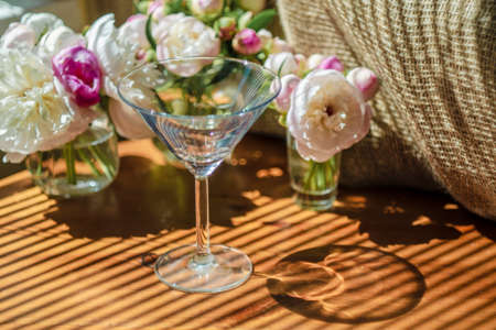 Festive rustic still life with glass empty clear glass surrounded by white flowers on textile background on wooden table. Diagonal striped shadow of the blinds on bright sunny day. Greeting card.の写真素材