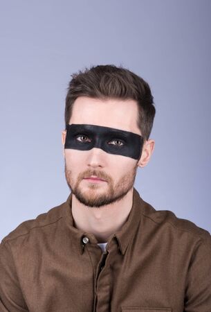 Portrait of a young male in a shirt with a drawn on face with black mask. Stylish hair and beard. Neutral view. Grey backgroundの写真素材