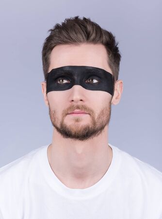 Portrait of a man in a white t-shirt. Black stripe on the eyes, stylish hair. Beard and mustache. Neutral grey backgroundの写真素材