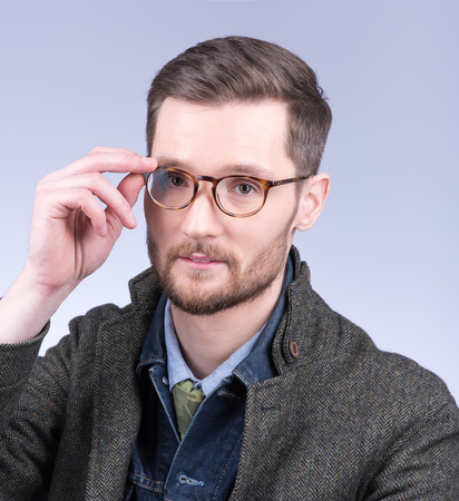 A young, stylish man with a beard adjusts her glasses. Modern dress, studio portrait. Neutral grey backgroundの写真素材