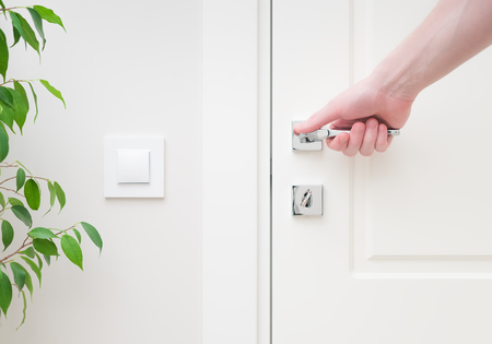 Male hand holding modern door handle. Close-up elements of the interior of the apartment. White door and lock with key. The light switch on the wall and green plant leavesの写真素材