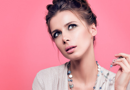 Horizontal closeup portrait. Young beautiful woman holds beads and dreamy looks up or chooses something. Gentle style, hair style and light makeup. Bright blouse. Pink backgroundの写真素材