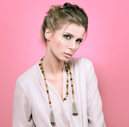 A delicate portrait in pink colors. Beautiful young woman in a light blouse on a pink background. Beads or decoration on the neck. Hairstyle and professional makeup. Gentle way, slightly lowered headの写真素材