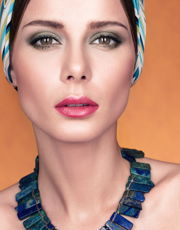 Beauty closeup portrait of beautiful young woman with kerchief on her head. Necklace on her neck. Makeup and fresh skin. Golden backgroundの写真素材