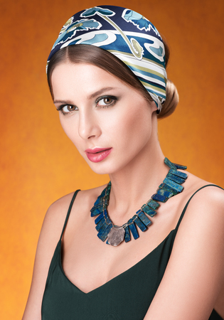 Hairstyle with headscarf. Portrait of young beautiful woman with necklace on her neck. Golden-red neutral backgroundの写真素材