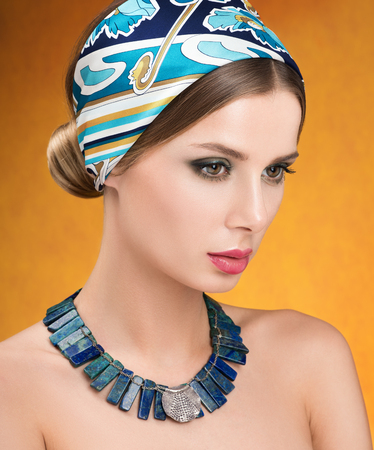 Necklace and headscarf. Beauty portrait of young beautiful woman, the gaze directed downwards. Contrasting makeup. Golden backgroundの写真素材
