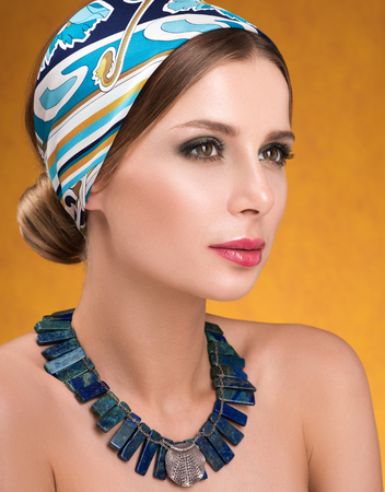 Hairstyle with headscarf. Closeup portrait of young woman with necklace on her neck. Golden-red neutral backgroundの写真素材