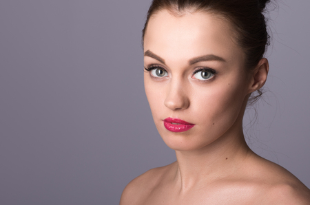 Closeup portrait of a beautiful young lady with clean, perfect skin. Professional light makeup and red lips. Shoulders bare, the hair gathered on the head. Neutral grey backgroundの写真素材