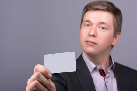 Horizontal closeup portrait of young attractive man in a jacket and shirt showing your business card or any other card. Neutral grey backgroundの写真素材