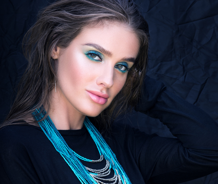 Closeup portrait of beautiful young woman in black. Professional makeup and hairstyle. Turquoise necklace on her neck. Black clothes and black backgroundの写真素材