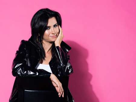 Beautiful young woman sitting on a chair and support your head with your hand. Pink background with free space. Stylish appearance, ripped jeans and a black jacketの写真素材