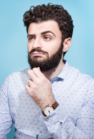 Partret brooding young men. Dark hair, beard and mustache. Light shirt and wrist watch. Neutral blue backgroundの写真素材