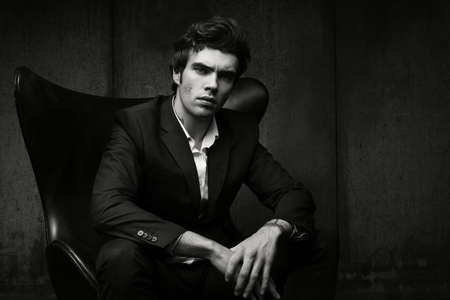 Stylish young man sitting in a chair. Black jacket and white shirt. Black-and-white characteristic portraitの写真素材