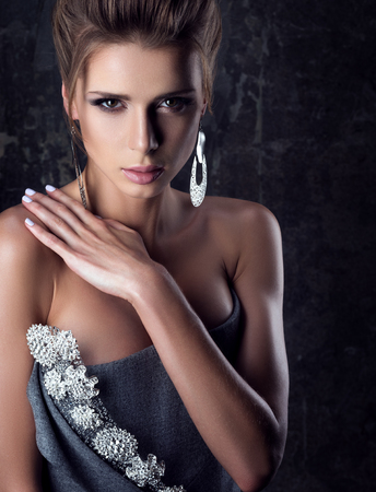 Portrait of young beautiful woman with sensual look. Professional makeup and large earrings. The hand on the shoulder. Evening dress with bare shoulders. The dark backgroundの写真素材