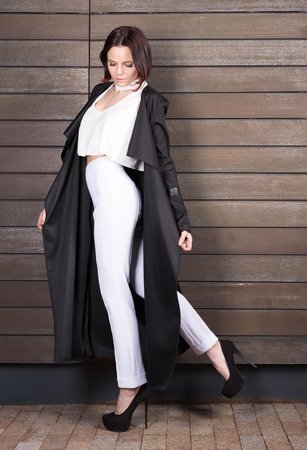 Young stylish woman in a black cloak and white pants posing on the background of wooden wallの写真素材