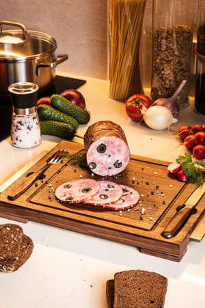 Smoked ham stuffed with black olives on a wooden board. Fresh vegetables and bread on the kitchen table around.の写真素材