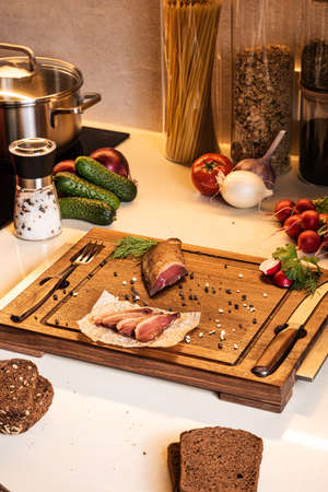 Smoked ham on a wooden board among vegetables and bread on the kitchen table. Fresh breakfast ingredients.の写真素材