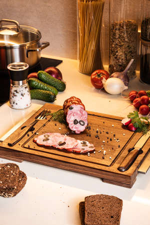 Cured sausage stuffed with black olives on a wooden board among vegetables and bread on the kitchen table.の写真素材