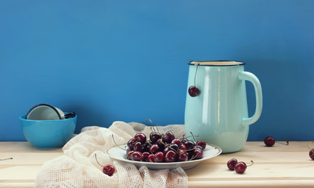 Still life with cherry and enameled jug on the table on a blue background.  Ripe berries in a plate.の写真素材
