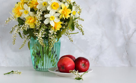A colorful arrangement of yellow daffodils and white flowers is placed in a green vase, besides three fresh red apples on a white plate, enhancing the fresh and cheerful atmosphere of spring.の写真素材