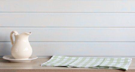 White ceramic pitcher and folded green checkered tablecloth resting on wooden table against white wooden wallの写真素材