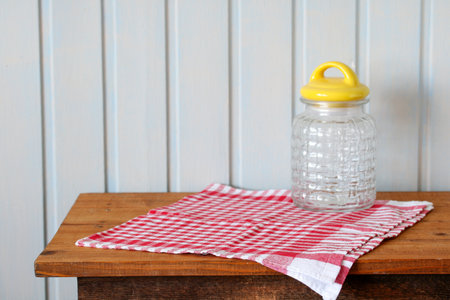 A transparent glass jar with a yellow lid is set on a red and white checkered cloth, resting on a rustic wooden table. The uncluttered, light blue panel background accentuates the simplicity of the arrangement.の写真素材
