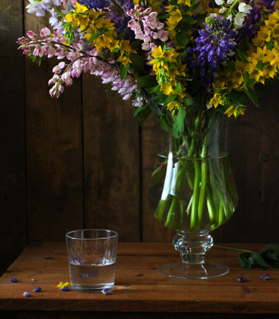 A colorful arrangement of wildflowers in a clear vase sits gracefully on a wooden table, accompanied by a glass of water. Soft evening light enhances the natural beauty of the blossoms.の写真素材