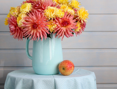 A stunning arrangement of pink and yellow flowers sits in a blue jug on a light blue table. Next to the jug, a fresh apple adds a touch of color, enhancing the overall charm of the setting.の写真素材