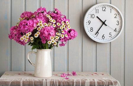 Pink and white flowers arranged in white pitcher next to clock on wallの写真素材