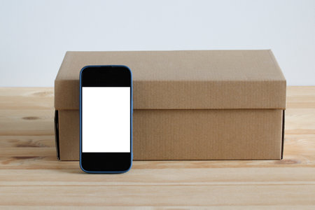 Smartphone with blank white screen leaning on closed cardboard box on wooden tableの写真素材