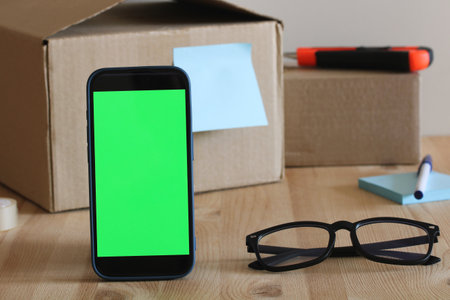Mobile phone with chroma key screen standing on desk with cardboard boxes, glasses and office supplies, perfect for showcasing apps or websitesの写真素材